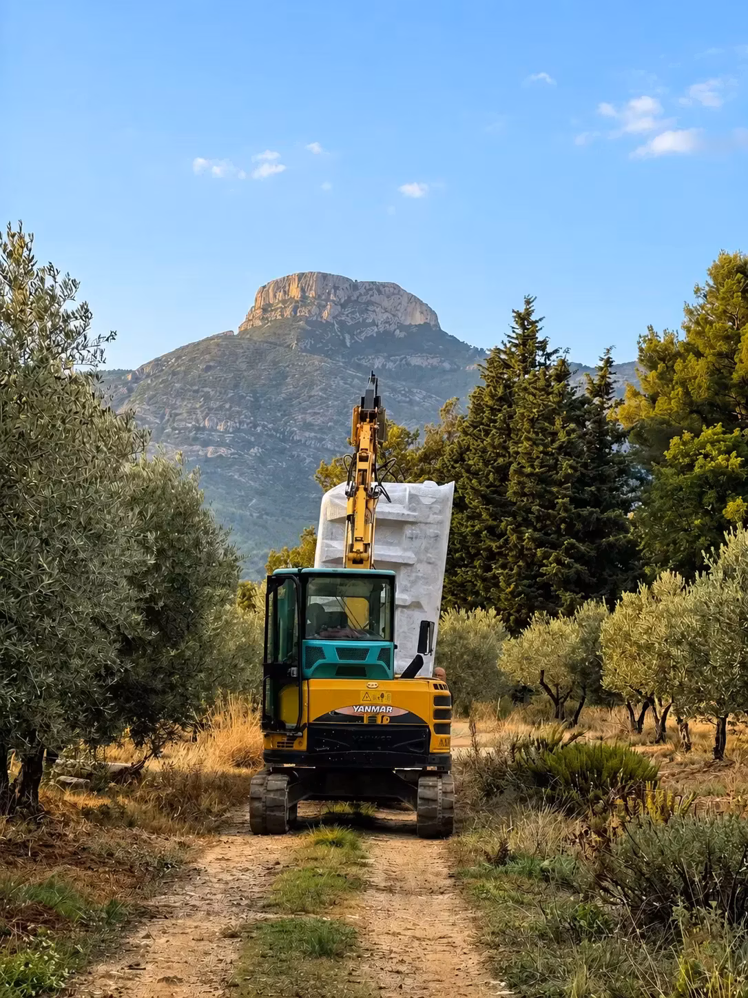 Pelleteuse jaune sur un chemin de terre menant à un chantier de piscine à Aubagne, oliviers et montagne en arrière-plan