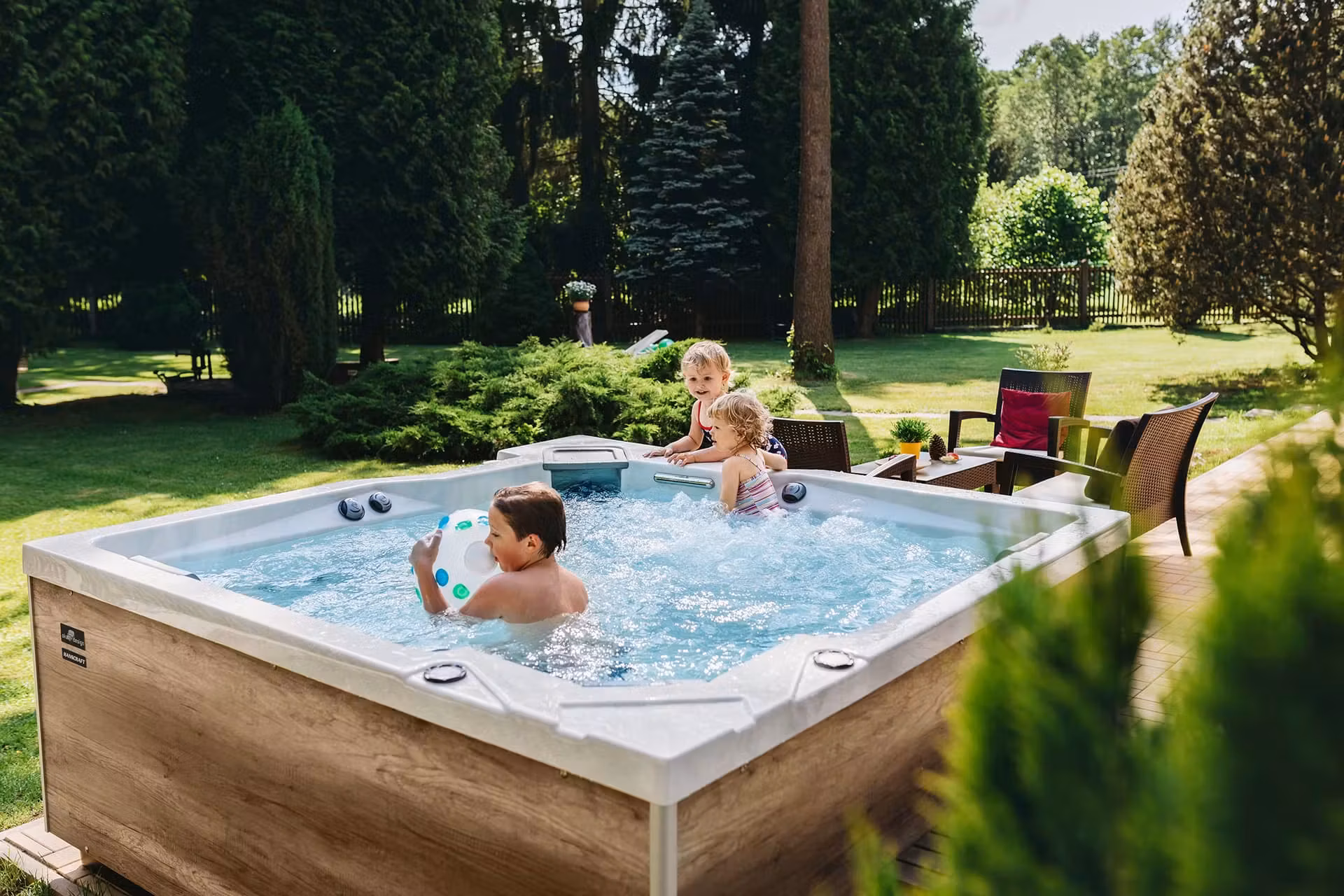 Trois enfants dans un spa Hanscraft installé en jardin, habillage bois clair, jeu avec un ballon