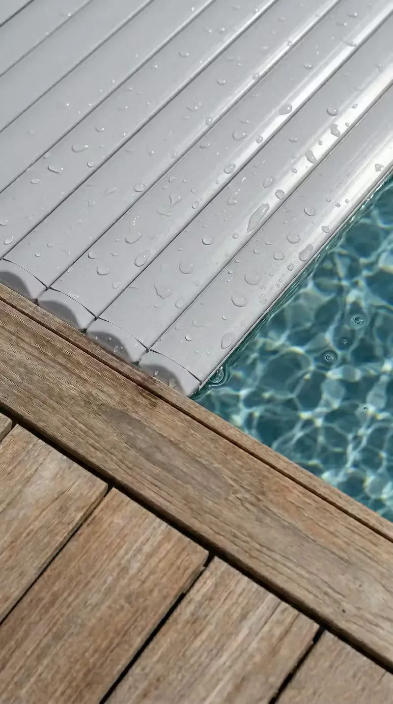 Détail d'un volet roulant immergé refermé sur une piscine, gouttes d'eau apparentes et margelle en bois