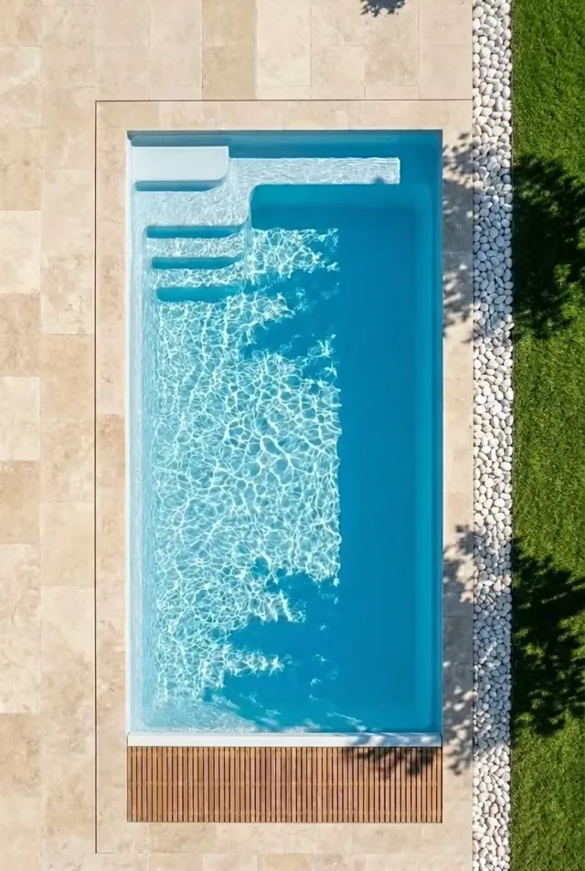 Vue aérienne d'une piscine coque avec volet immergé, margelles en travertin et bordure de galets blancs