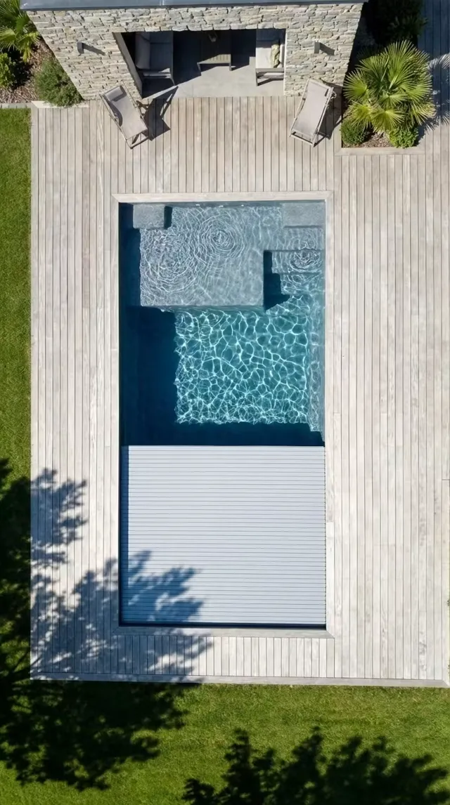Vue aérienne d'une piscine coque avec volet roulant immergé en cours de fermeture, deck en bois clair et pool house en pierre